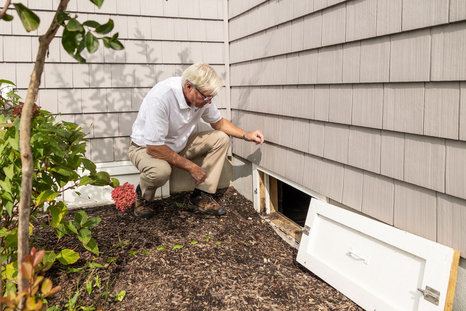 Home inspector crouched beside a house with the crawl space access door open, checking the space beneath the floor