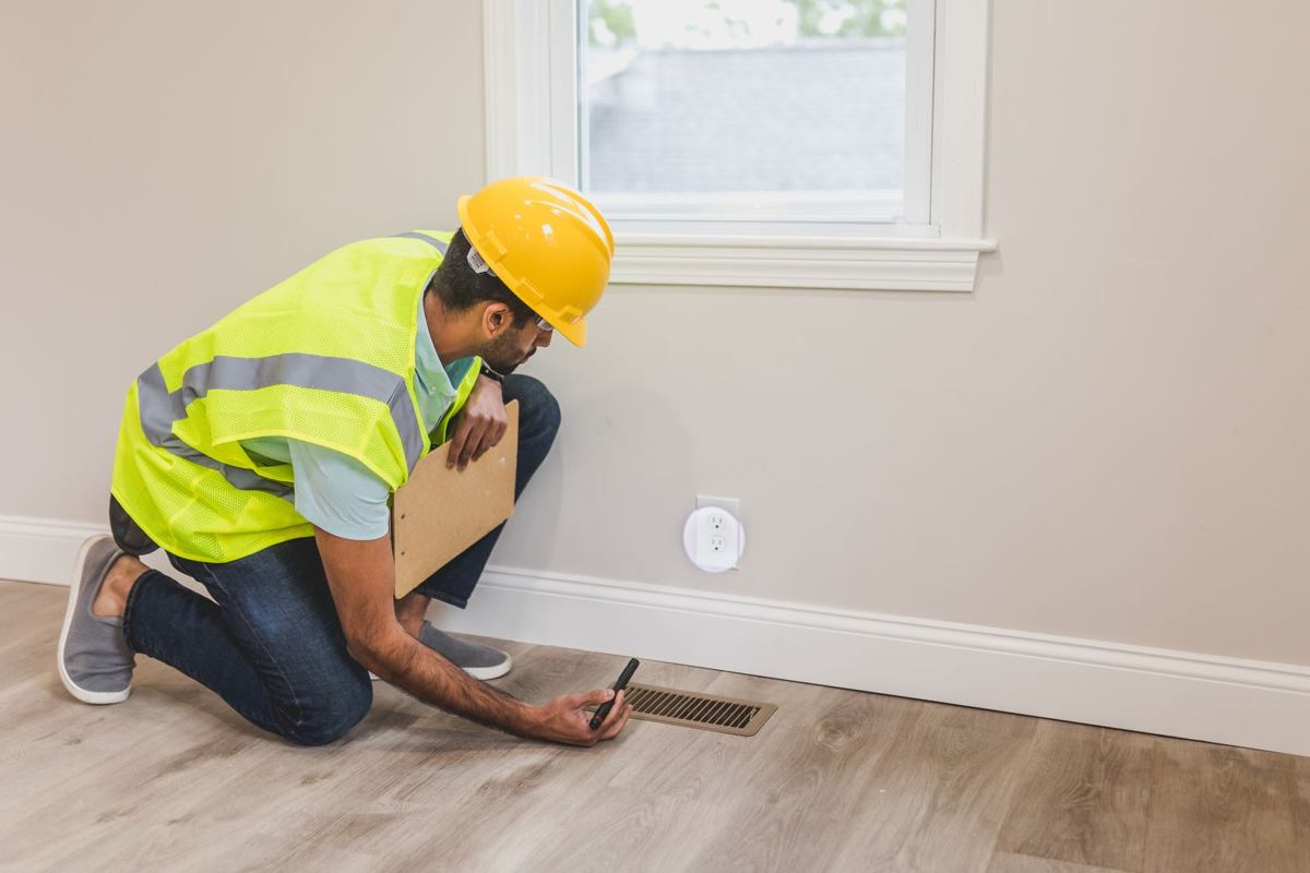 Inspector in a safety vest kneeling on a hardwood floor, checking a floor register where crawl space air enters the home